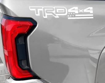 May include: A close-up of a silver vehicle's rear panel, featuring a white "TRD 4x4" decal. The decal includes the text "TRD" in a bold font, followed by "4x4" and a stylized American flag design. The vehicle's red taillight is visible.