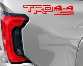 May include: Red TRD 4x4 decal on a silver vehicle. The decal features the letters "TRD" in a bold font, followed by "4x4" and a graphic of the American flag. The vehicle's red taillight is visible on the left.