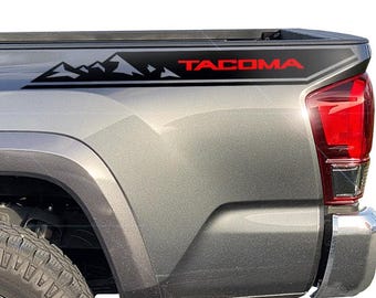 May include: A gray pickup truck with a black and red graphic on the side. The graphic features a mountain range and the word "TACOMA" in red. The truck's taillight is visible.