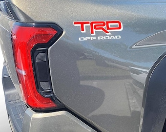 May include: A gray pickup truck with a red "TRD OFF ROAD" emblem on the back.