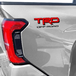 May include: A silver pickup truck with a red "TRD OFF ROAD" emblem on the back.
