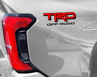 May include: A silver pickup truck with a red "TRD OFF ROAD" emblem on the back.