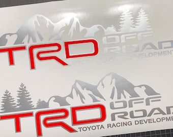May include: Two white decals with the text "TRD OFF ROAD TOYOTA RACING DEVELOPMENT". The letters "TRD" are red with a silver outline. The decals feature a mountain range and pine trees in silver.