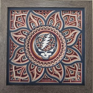 May include: A layered paper art piece featuring a skull with a lightning bolt on a mandala design. The artwork uses shades of red, cream, and blue, framed in a gray wooden frame. The design is intricate and detailed.