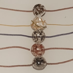 May include: Close-up of five decorative beads on strings. The beads are clear with speckled patterns in brown, gold, and black. The strings are in various colors, including brown, yellow, and blue. The beads are evenly spaced.