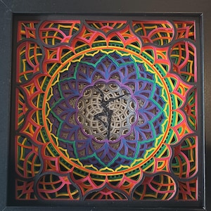 May include: A layered paper art piece in a black frame. The artwork features a vibrant, multi-colored mandala design with a central silhouette of a dancer. The colors include red, orange, yellow, green, purple, and blue.
