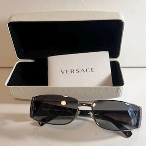 May include: Dark grey rectangular sunglasses with a metallic frame and a Greek key pattern on the temples. The sunglasses are positioned in front of an open white hard-shell case with a black interior, containing a white card printed with VERSACE.