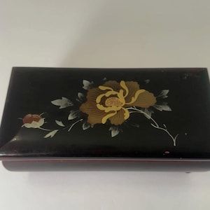 May include: A rectangular, black wooden jewelry box with a floral design. The lid features a large, gold and brown flower with white leaves and a smaller red and gold flower. The box has a glossy finish and a red base.