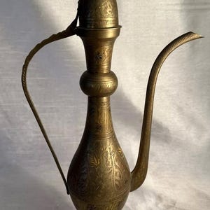 Puede incluir: Una tetera de latón de color dorado con un diseño ornamentado. La tetera tiene un pico largo y curvo y un asa que está unida a la tapa. La tapa tiene un remate en la parte superior.