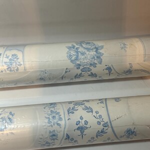 May include: Two rolls of wallpaper wrapped in clear plastic, featuring a repeating pattern of blue floral bouquets and intricate botanical vines set against a cream-colored background, with vertical stripes separating the floral design elements.