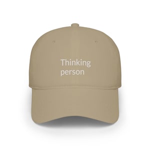 Può includere: Un cappellino da baseball color marrone chiaro con corona strutturata e visiera curva. Il pannello frontale presenta la scritta "Thinking person" stampata in un colore bianco sporco chiaro. Il cappellino include un bottone e occhielli.