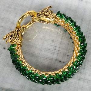 May include: A circular bracelet featuring a gold-toned metal dragon head clasp and a band constructed from small, interlocking green and gold beads. The design mimics the appearance of scales, with the dragon heads meeting at the top closure.