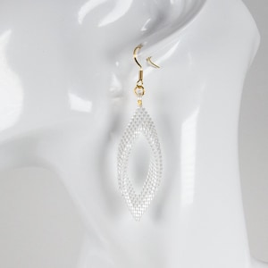 May include: A white beaded earring with a diamond-shaped open center, suspended from a gold-toned metal hook. The earring features a fine, intricate mesh-like texture. The logo BEADED DRIP is visible in the upper right corner of the image.