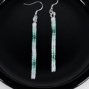 May include: A pair of long, slender drop earrings featuring a tubular design crafted from tiny beads in a gradient pattern of white, light green, and dark green. The earrings hang from silver-toned metal hooks and include the text BEADED DRIP.