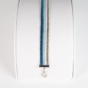 May include: A beaded bracelet with a gradient of blue, silver, and white beads. The bracelet is displayed on a white cylindrical stand. The clasp is silver and has a small charm. The logo "Beaded Drip" is in the upper left corner.