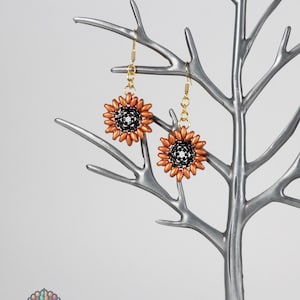 May include: Sunflower-shaped beaded earrings with copper-colored petals and a black center, hanging from gold-colored ear wires. The earrings are displayed on a silver-colored decorative tree with a small bird perched on a branch.