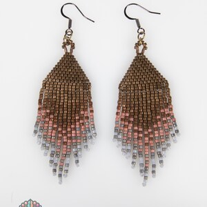 May include: A pair of dangle earrings featuring a woven bead design that transitions from a metallic bronze at the top to a soft pink and light gray at the bottom fringe. The earrings are suspended from dark metal hooks and include a logo.