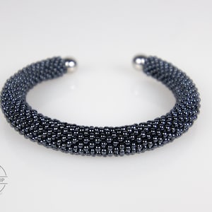 May include: A dark navy blue beaded cuff bracelet featuring a dense, uniform texture of small, reflective spherical beads. The bracelet has a circular, open-ended design with smooth, polished silver-toned metal end caps at each terminal point.
