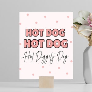 May include: Pink and white printable sign with the text "HOT DOG HOT DOG Hot Diggity Dog" in black and pink lettering. The background has a pink polka dot pattern.