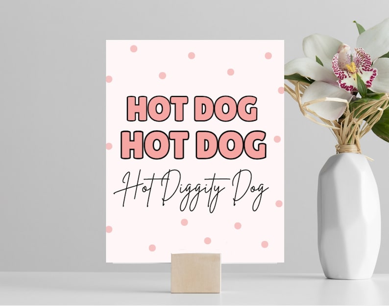 May include: Pink and white printable sign with the text "HOT DOG HOT DOG Hot Diggity Dog" in black and pink lettering. The background has a pink polka dot pattern.