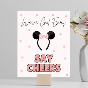 May include: A white sign with pink polka dots and the text "We've Got Ears Say Cheers" in black and pink. A black headband with pink ears and a pink bow is in the center of the sign.