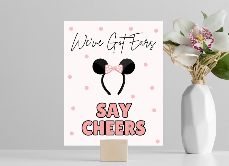 May include: A white sign with pink polka dots and the text "We've Got Ears Say Cheers" in black and pink. A black headband with pink ears and a pink bow is in the center of the sign.