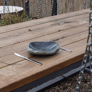 Può includere: Una scena di un tavolo di legno rustico all'aperto con una ciotola di pietra, una forchetta e un coltello. La ciotola ha un aspetto scuro e marmorizzato. Una catena di metallo è appesa sullo sfondo.