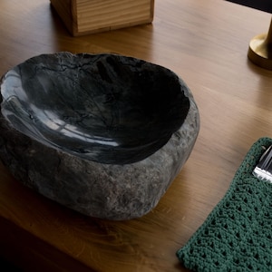 Pode incluir: Uma tigela cinza escuro, semelhante a pedra, com um interior liso está em uma superfície de madeira. Um porta-utensílios verde de crochê com talheres está à direita. Um objeto dourado e uma caixa de madeira estão ao fundo.