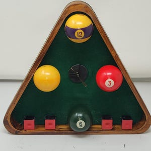 Könnte beinhalten: Eine dreieckige Uhr aus Holz mit grünem Filzinnenraum, die Billardkugeln mit den Nummern 3, 6 und 12 zeigt. Die Uhr hat einen dunklen Holzrahmen und kleine rote quadratische Akzente an der Basis.