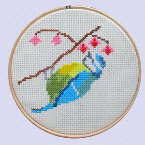 May include: Cross-stitch embroidery hoop depicting a blue tit perched on a branch with pink blossom. The bird is blue, green and yellow with a white breast.