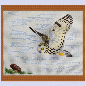 May include: A cross-stitch embroidery depicting a bird in flight, with a small mouse in the bottom left corner. The bird is white, grey, and yellow with black markings. The background is a light blue with white lines.