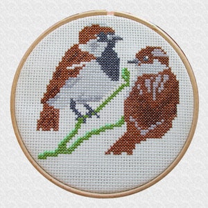 May include: Cross-stitch embroidery hoop featuring two brown birds perched on green stems. The birds are stitched in a detailed style using brown, white, and grey thread.