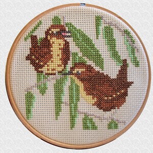 May include: A cross-stitch embroidery depicting two brown birds perched on green branches. The birds are surrounded by green leaves and are stitched in a realistic style.