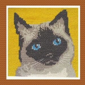 May include: Cross-stitch depiction of a Siamese cat, featuring vivid blue eyes and dark facial markings. The cat's fur is a blend of grey and white, set against a yellow backdrop. The artwork is framed with a white border, showcasing the detailed needlework.