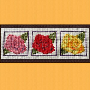 May include: A cross-stitch floral design with three square panels. Each panel displays a rose in shades of pink, red, and yellow, with green leaves. The artwork is framed in a dark brown border against an orange background.
