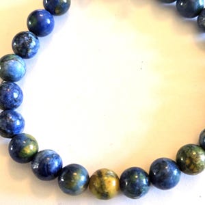 May include: A beaded bracelet featuring round, polished beads in shades of blue, green, and yellow. The beads are strung together with a silver-colored clasp. The bracelet is displayed against a white background.