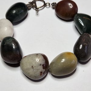 May include: A beaded bracelet featuring polished, oval-shaped stones in various colors, including black, white, brown, and green. The bracelet is secured with a silver-colored toggle clasp.