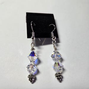 May include: A pair of dangling earrings with clear, faceted crystal beads and silver-colored metal accents. The earrings have a hook closure and are displayed on a black card against a white background. The crystals reflect multiple colors.