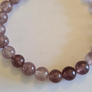 May include: A close-up of a bracelet featuring round, translucent beads in shades of purple and mauve. The beads have a marbled appearance and are strung together with a silver clasp.