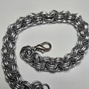 May include: A silver-colored chainmail bracelet with a lobster clasp. The bracelet is made of interlocking metal rings, creating a textured, woven appearance. The design is symmetrical and the clasp is attached to a small ring.