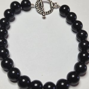 May include: A black beaded bracelet with a silver-colored toggle clasp. The bracelet is made of round, glossy black beads strung together. The clasp is a simple, elegant design.
