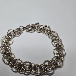 May include: A silver-colored chainmail bracelet with a toggle clasp. The bracelet is made of small, interlocking rings, creating a textured, flexible design. The clasp is a simple bar and ring closure.