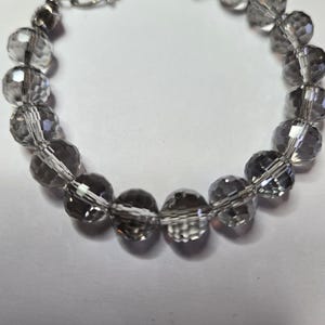 May include: A clear, faceted bead bracelet with a silver clasp. The beads are a smoky gray color and are strung together with a thin, silver-colored wire. The bracelet is designed to be worn around the wrist.