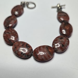 May include: A beaded bracelet featuring oval-shaped, reddish-brown stones with black veining. The bracelet is connected with a silver-colored clasp. The stones are smooth and polished, creating a natural and earthy aesthetic.