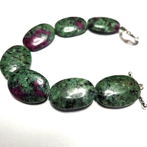 May include: A bracelet featuring oval-shaped beads of green zoisite with ruby inclusions. The polished stones are connected with a silver-colored clasp. The bracelet is displayed against a white background.