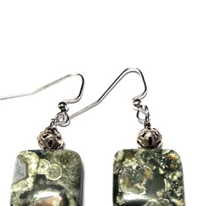May include: A pair of square earrings with a natural stone appearance. The earrings feature a silver-colored hook and a decorative silver bead above the square stone. The stone has a mix of green, gray, and white colors.