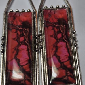 May include: A pair of rectangular earrings with a silver-colored metal frame. The earrings feature a red and pink marbled design encased in clear resin. The top of each earring has a loop for hanging.