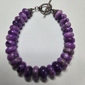 May include: A purple beaded bracelet with a silver toggle clasp. The beads are a mix of light and dark purple hues. The bracelet is made of round, polished beads. The clasp is a decorative silver design.
