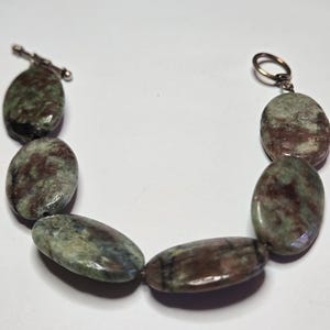 May include: A beaded bracelet featuring oval-shaped stones in shades of green, brown, and gray. The bracelet has a silver-colored toggle clasp closure. The stones are strung together, creating a unique accessory.