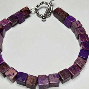 May include: A purple and brown square bead bracelet with a silver toggle clasp. The beads are a mix of dark purple and brown hues, creating a unique color pattern. The bracelet is designed to be worn around the wrist.